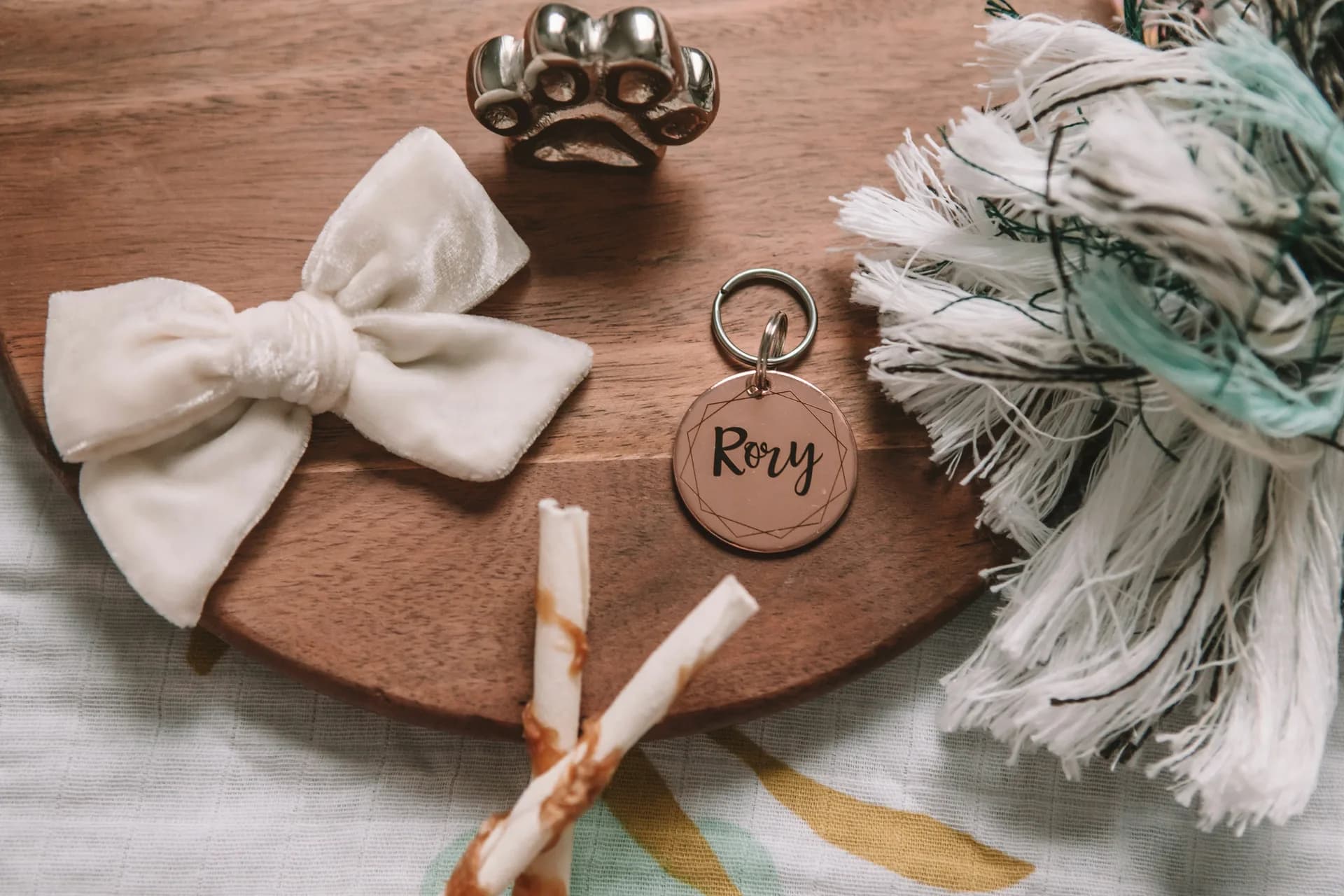 A personalized pet ID tag engraved with the name Rory beside a silver paw-print keepsake, a velvet bow, and a rope toy on a wooden board
