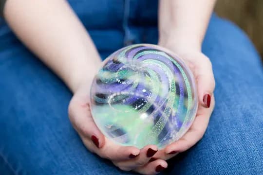 Hands cradling a handblown glass memorial orb with swirls of blue, green, and purple