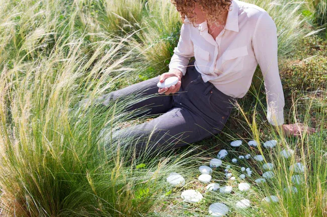 A person sitting in a sunlit meadow holding a solidified remains stone, with more pale stones resting in the grass around them.