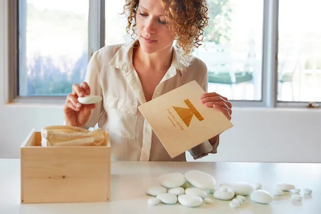 A woman lifting a smooth solidified remains stone from a wooden keepsake box on a table, with more stones spread in front of her.